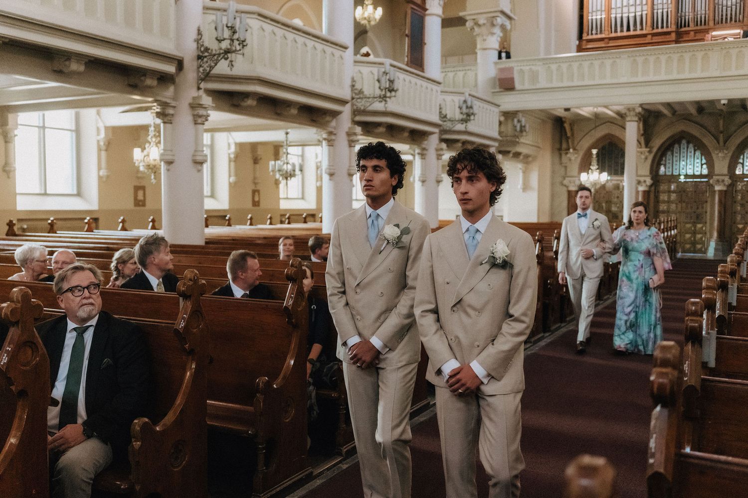 Two groomsmen in tan suits stand at church altar during wedding ceremony processional.