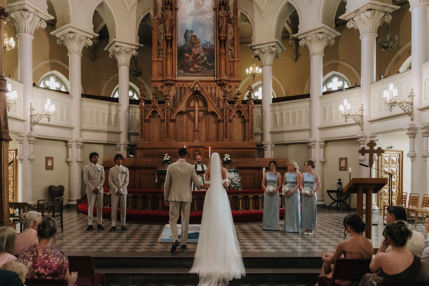 Wedding party stands at ornate church altar with bridesmaids in blue and groomsmen in tan suits.