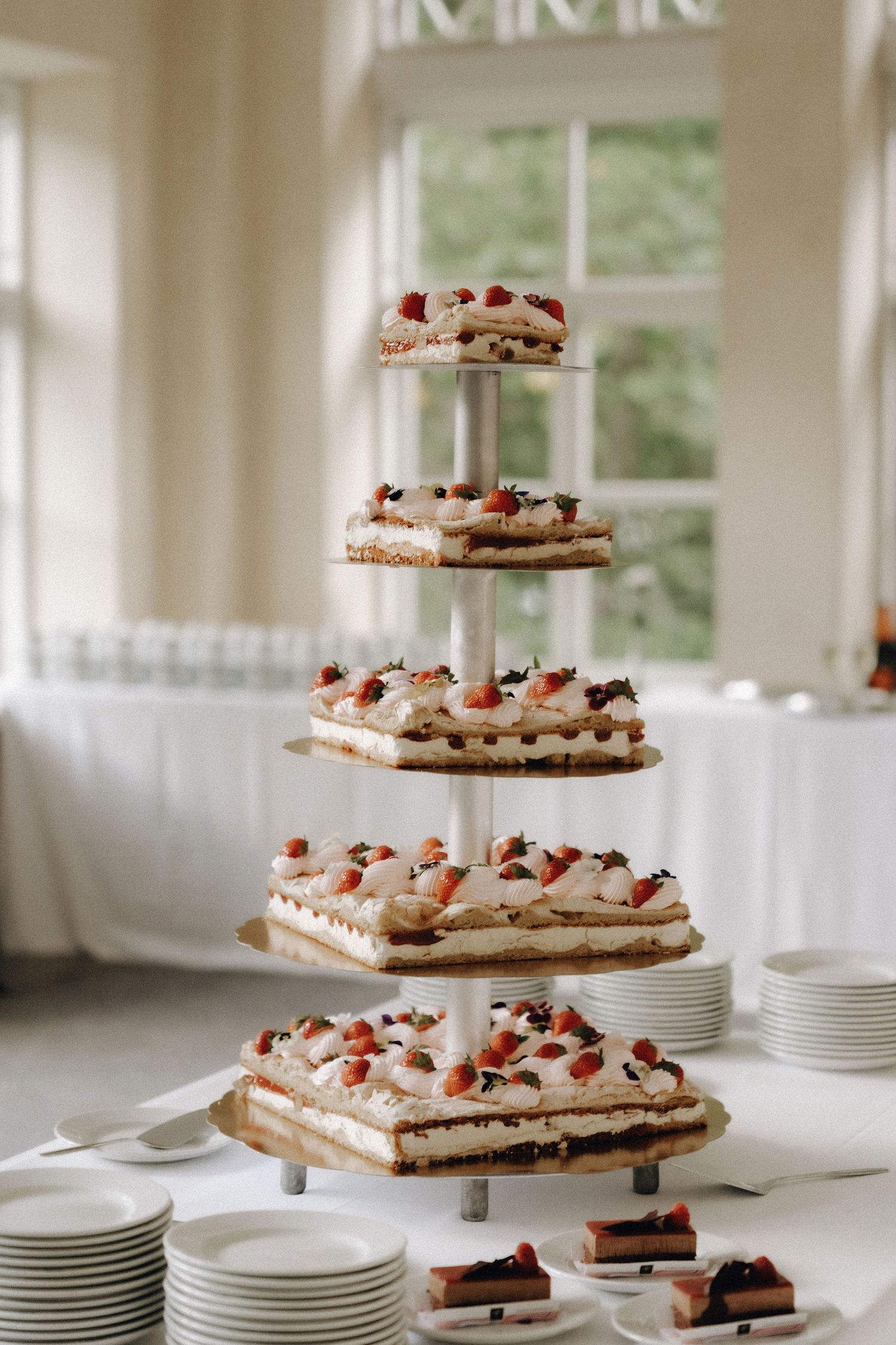 Five-tier wedding cake stand displaying square strawberry cream cakes near a bright window.