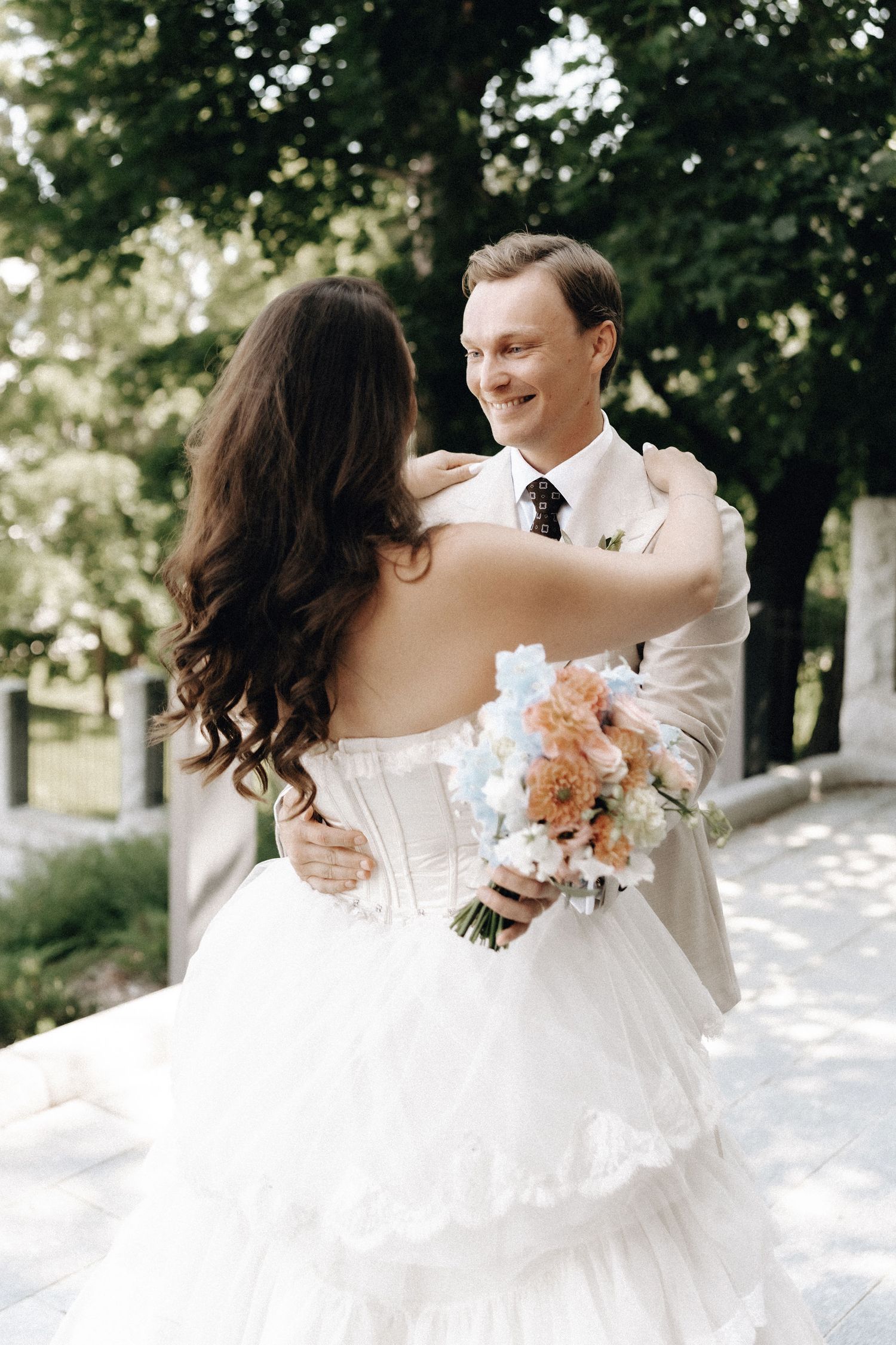 Smiling groom in beige suit embraces bride in strapless white ruffled gown holding pastel bouquet outdoors.