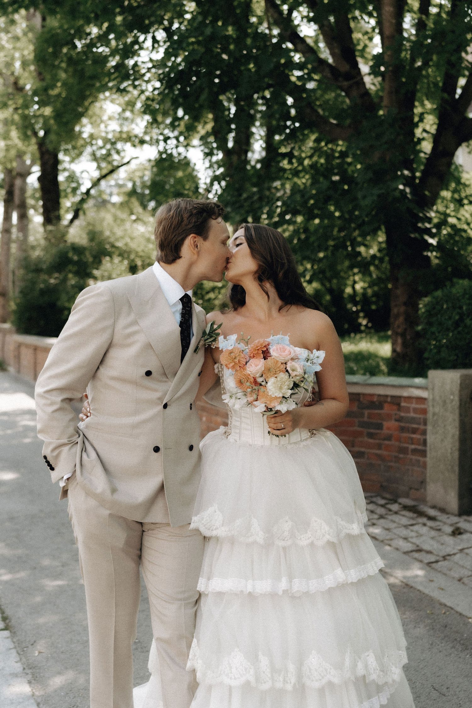 Bride and groom share a romantic kiss on a sunlit garden path, bride holding a colorful bouquet.
