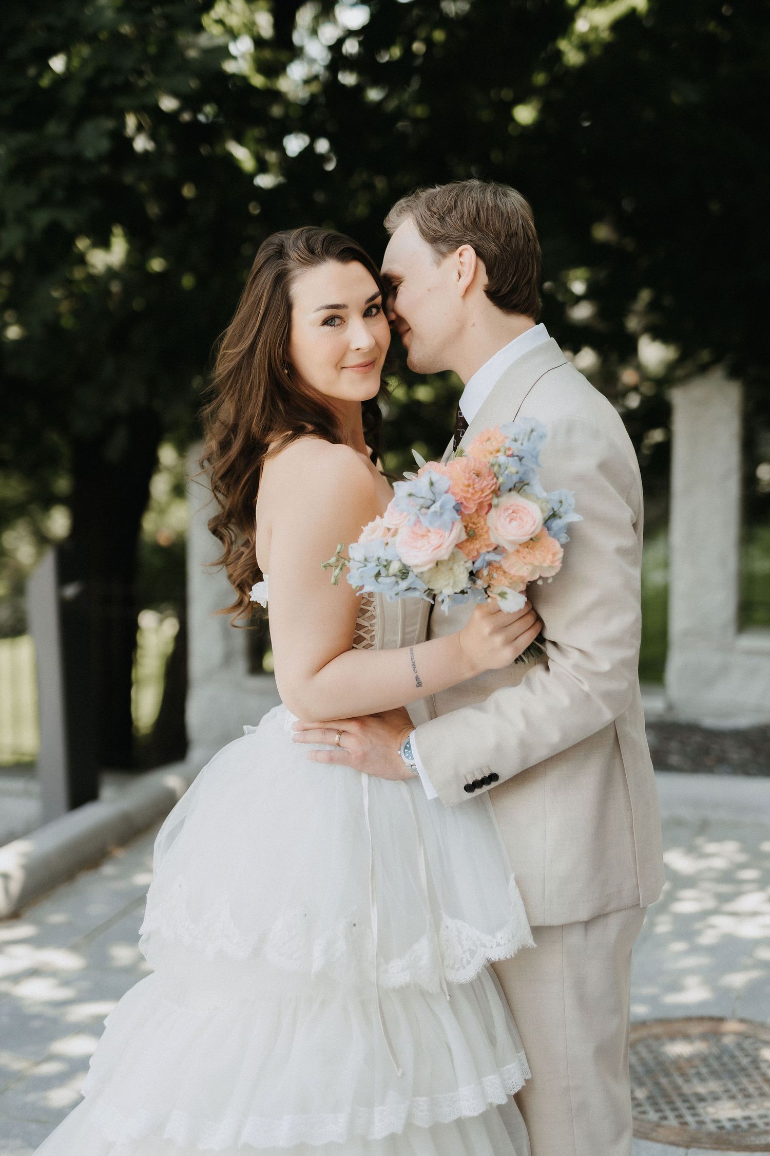 Bride in white ruffled gown smiles at camera while groom in beige suit whispers in her ear holding bouquet.