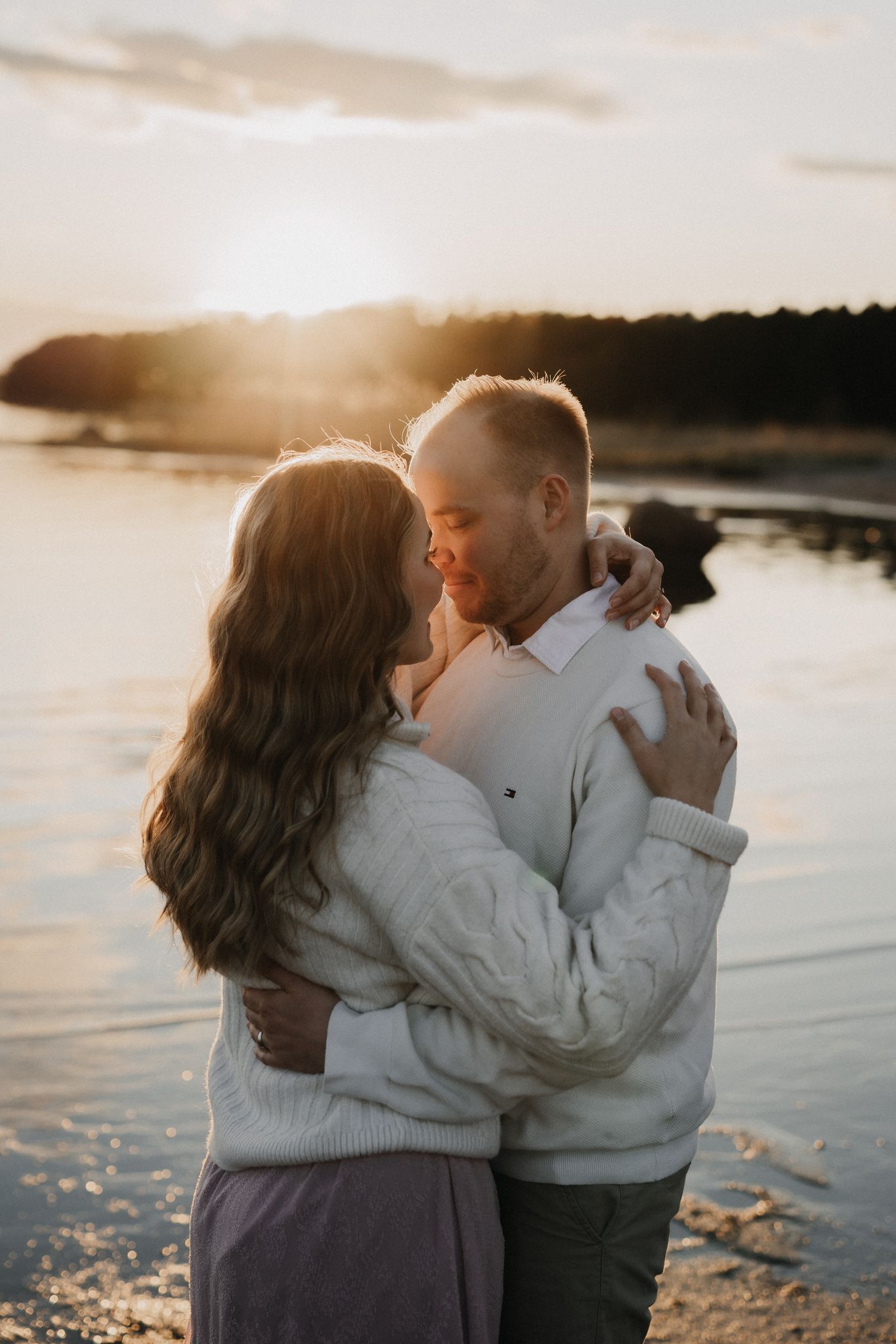 Couple shares a tender embrace on a beach at golden hour, backlit by a stunning sunset over the water.
