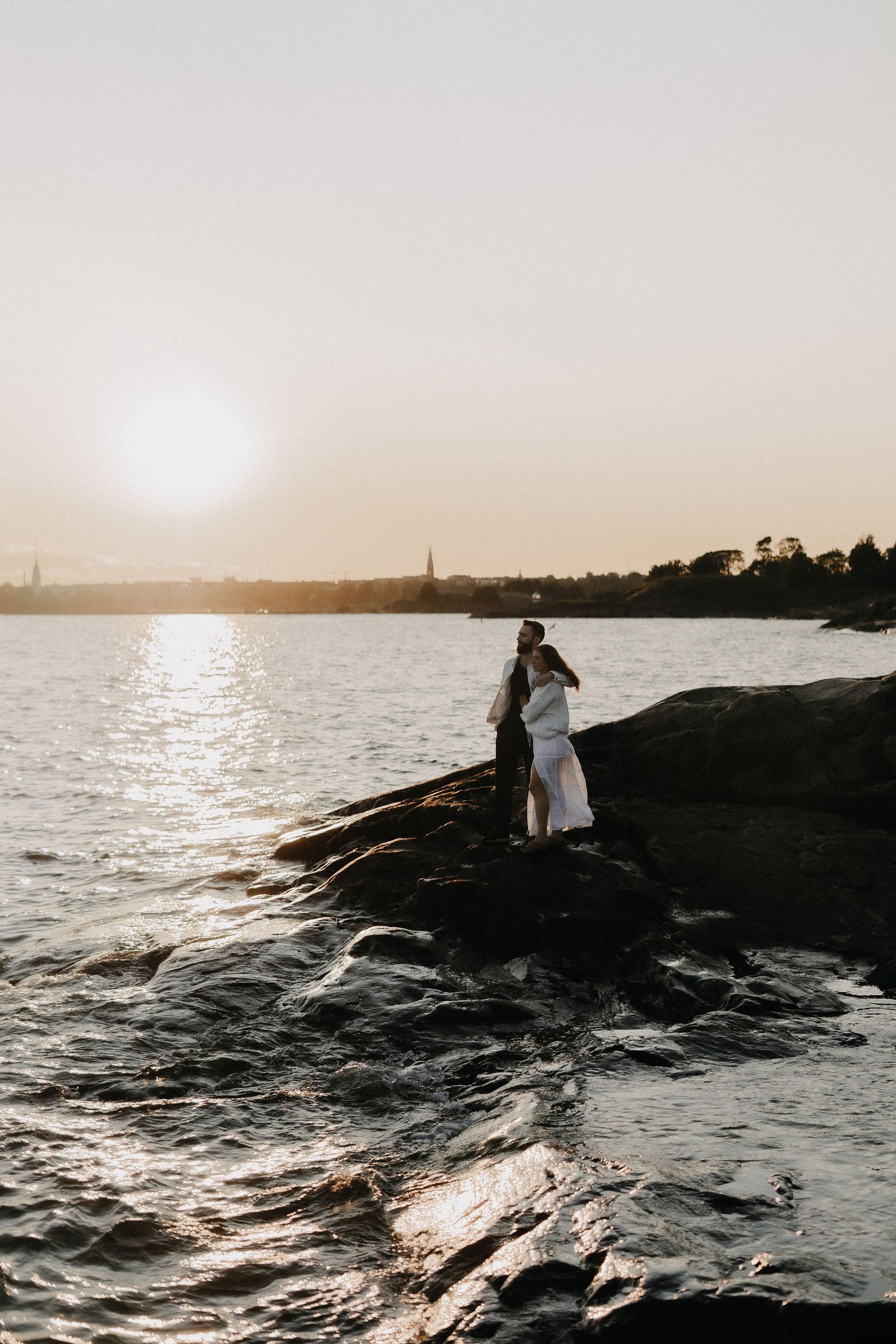 Romantic couple embracing on rocky shoreline at sunset with glowing water reflections behind them.