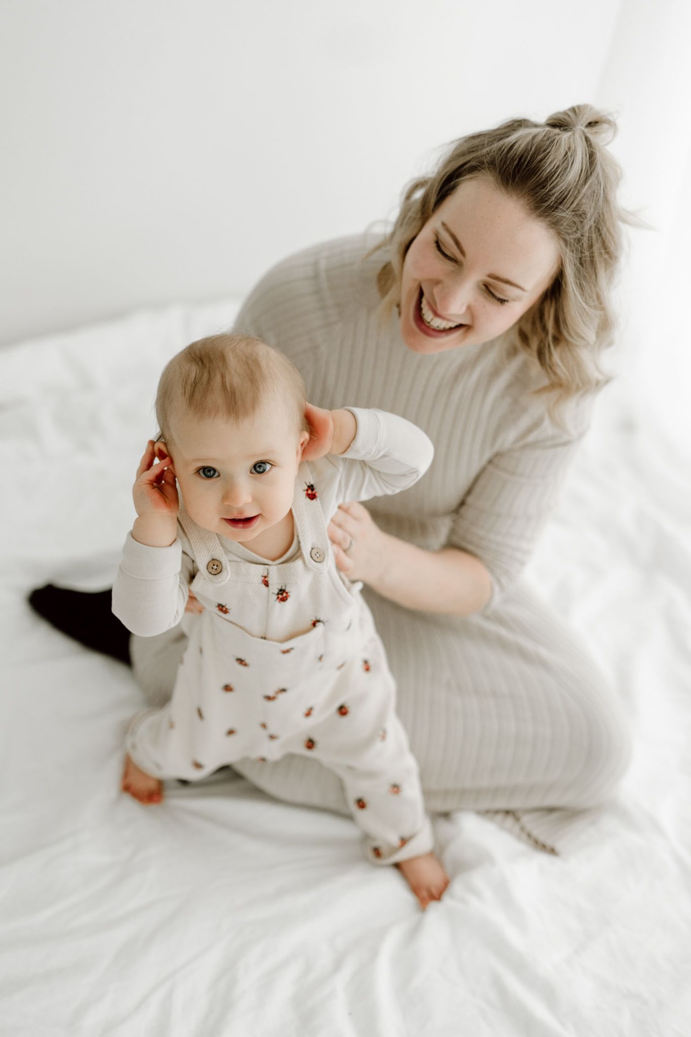 Smiling mother sitting on white bed helping baby in floral onesie stand, sharing a joyful moment.