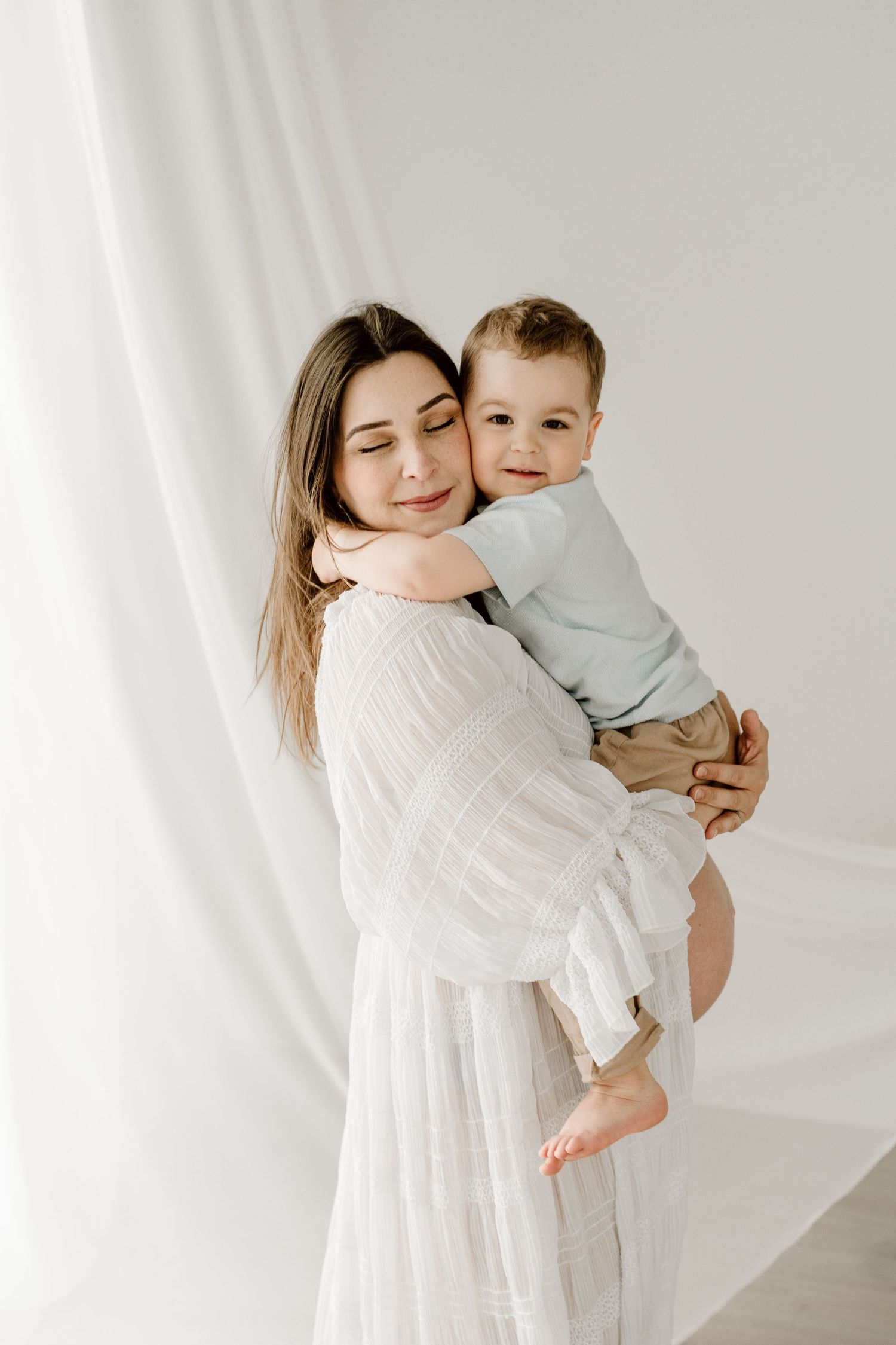 A pregnant woman in a white dress holds her smiling toddler son near flowing white curtains.