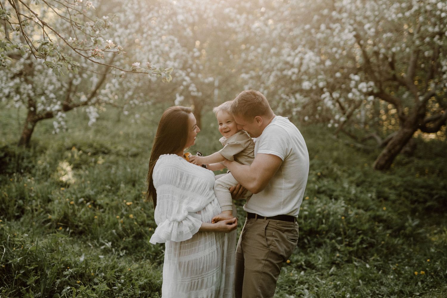 A happy family with a baby shares a tender moment among blossoming trees in a sunlit garden.