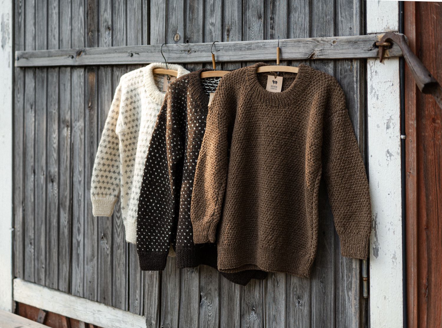 Three knitted sweaters in cream, grey, and brown hang on a rustic wooden barn wall display.