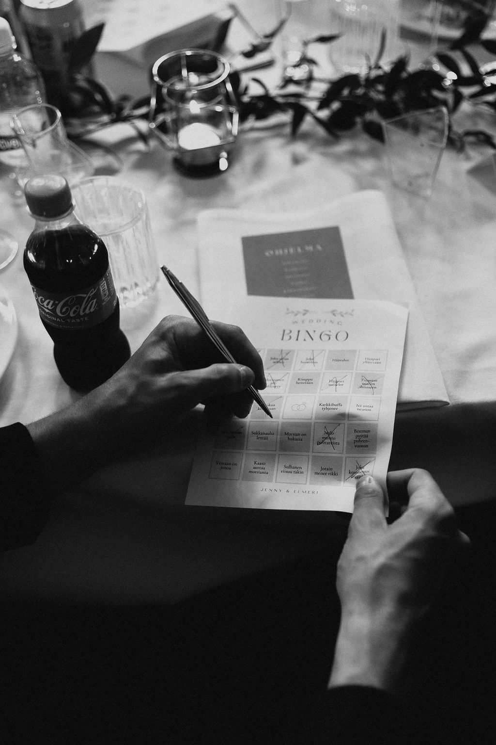 Wedding guest playing bridal bingo card game at reception table with Coca-Cola bottle nearby.