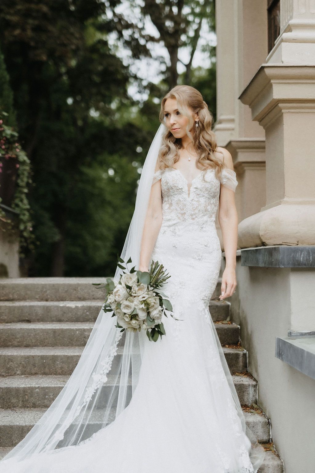 Bride in off-shoulder lace mermaid wedding dress with long veil holds bouquet on stone steps.