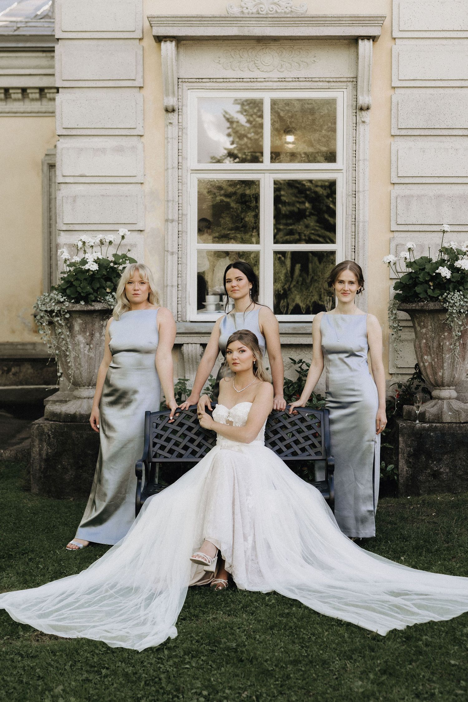 Bride in strapless ball gown seated with three bridesmaids in silver satin dresses at estate.
