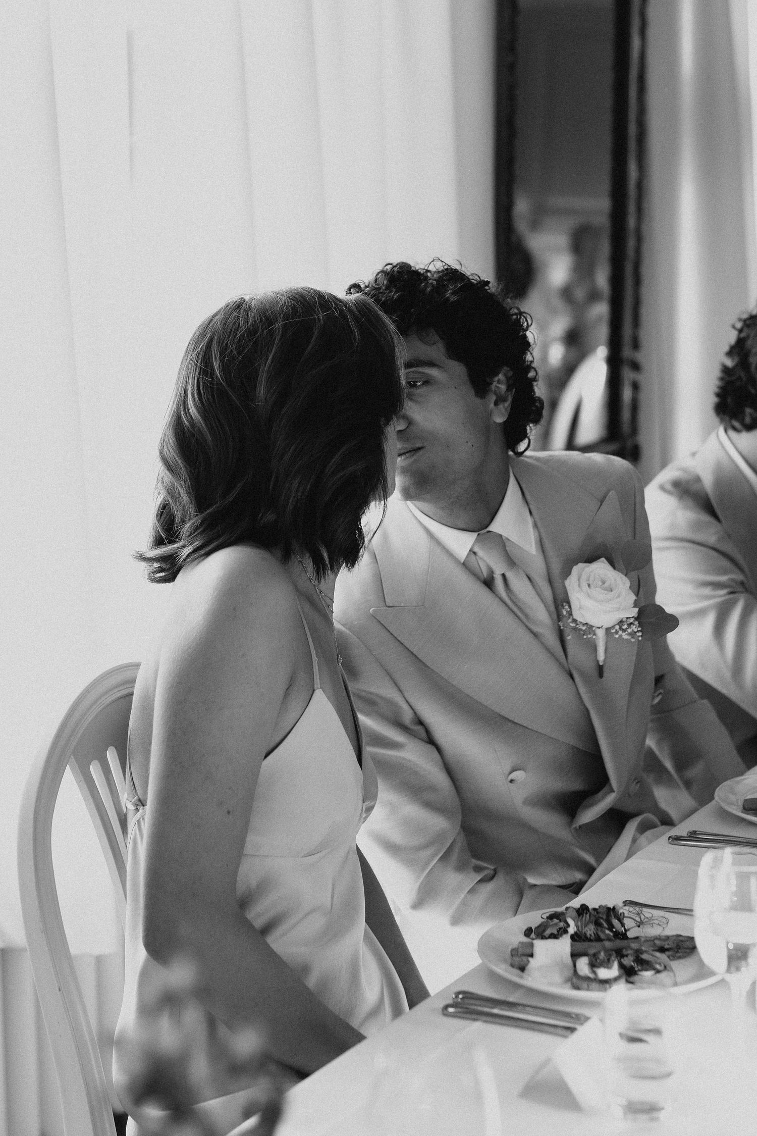 Black and white photo of groom in suit sharing an intimate moment with guest at wedding reception.