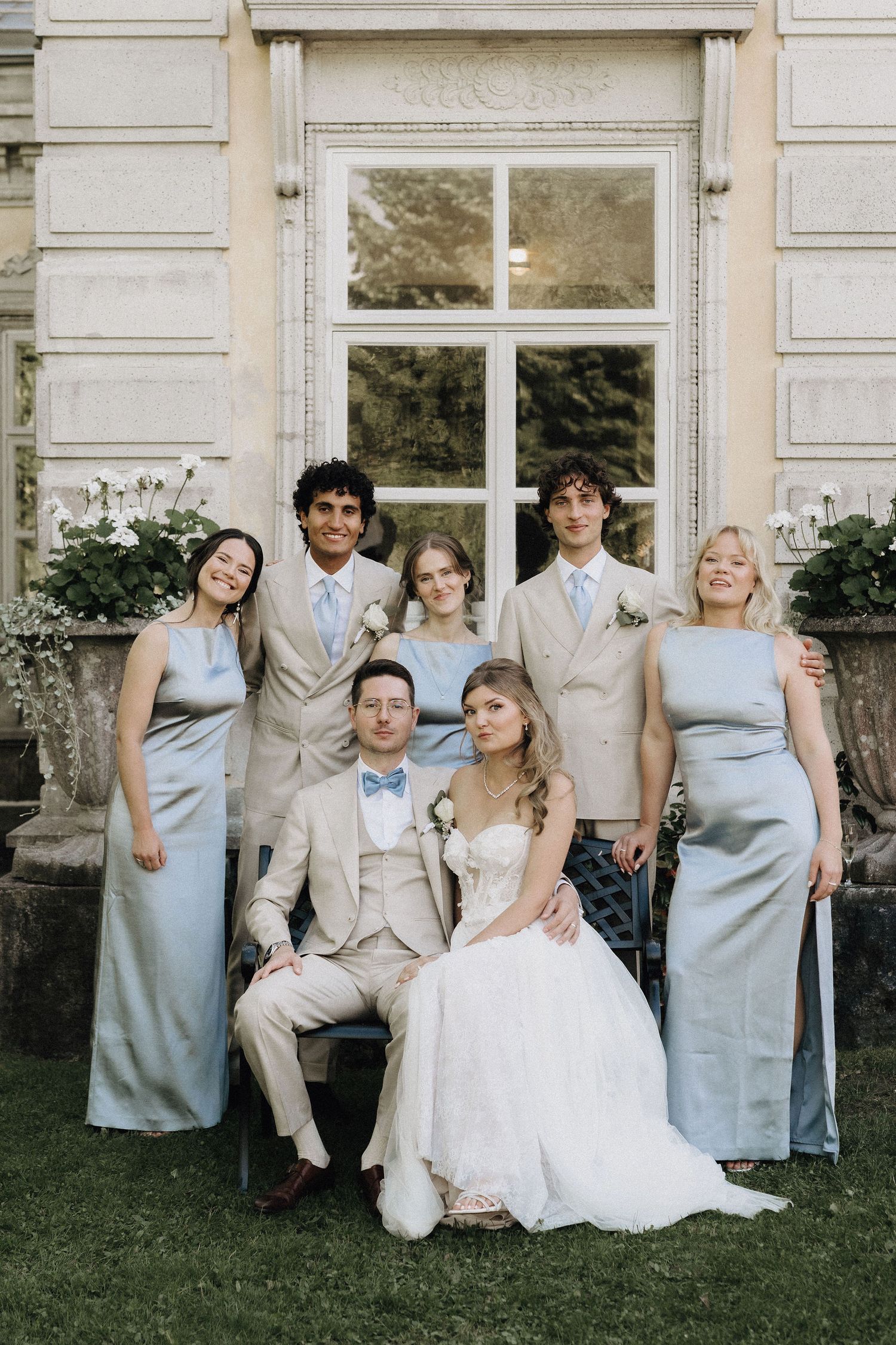 Wedding party portrait with bride, groom, bridesmaids in blue-grey satin and groomsmen in beige suits outdoors.