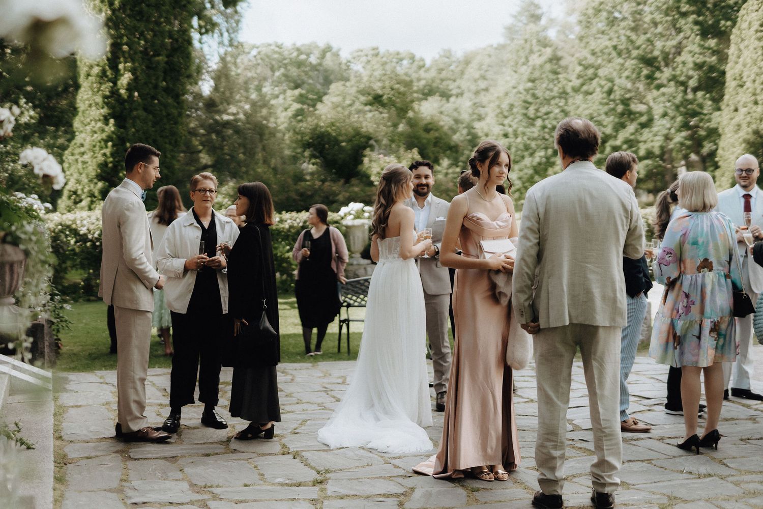Wedding guests mingle outdoors in a garden setting as the bride in white chats with attendees.