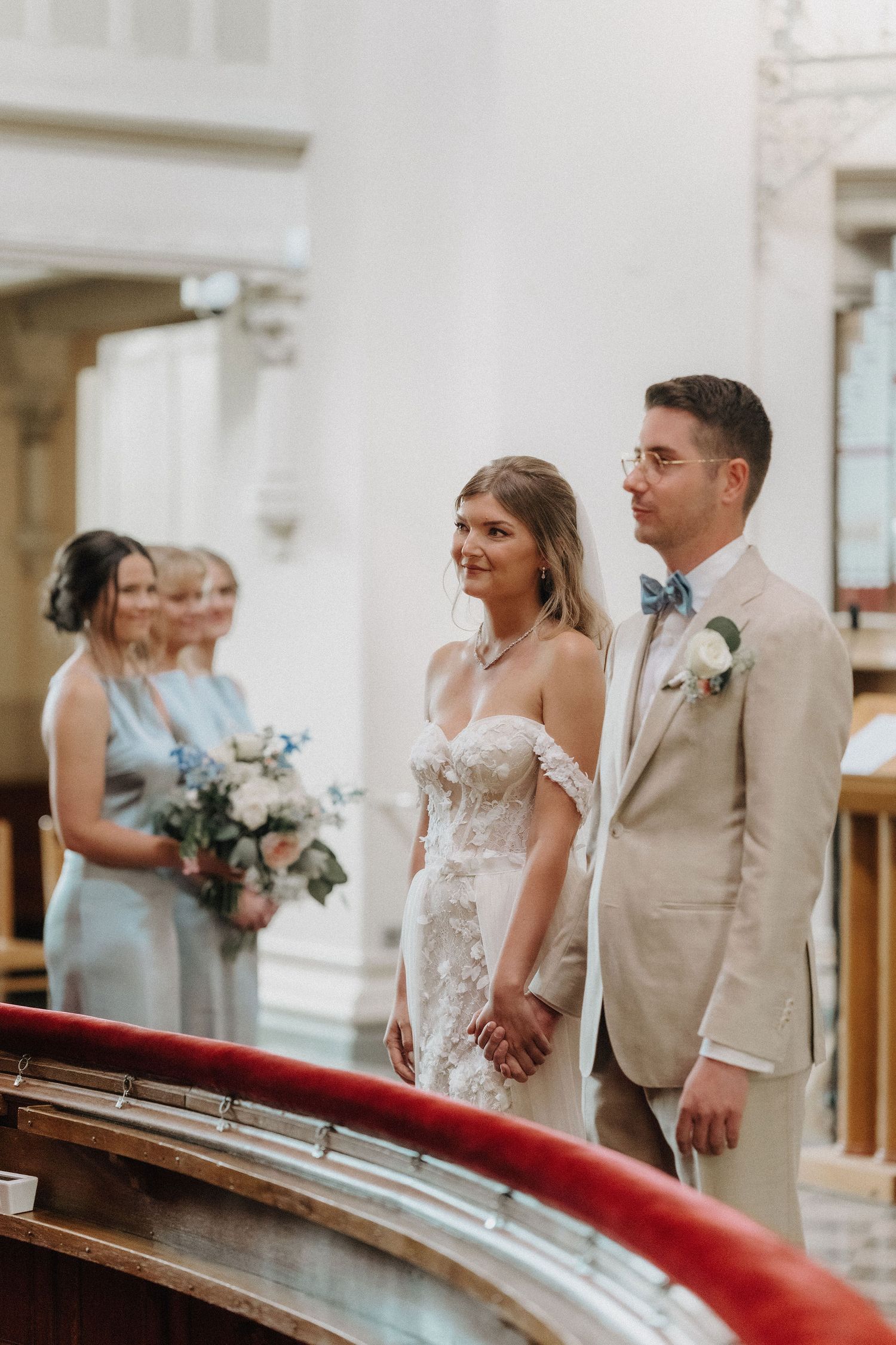 Bride in lace off-shoulder gown and groom in tan suit holding hands at the altar with bridesmaids in blue behind.