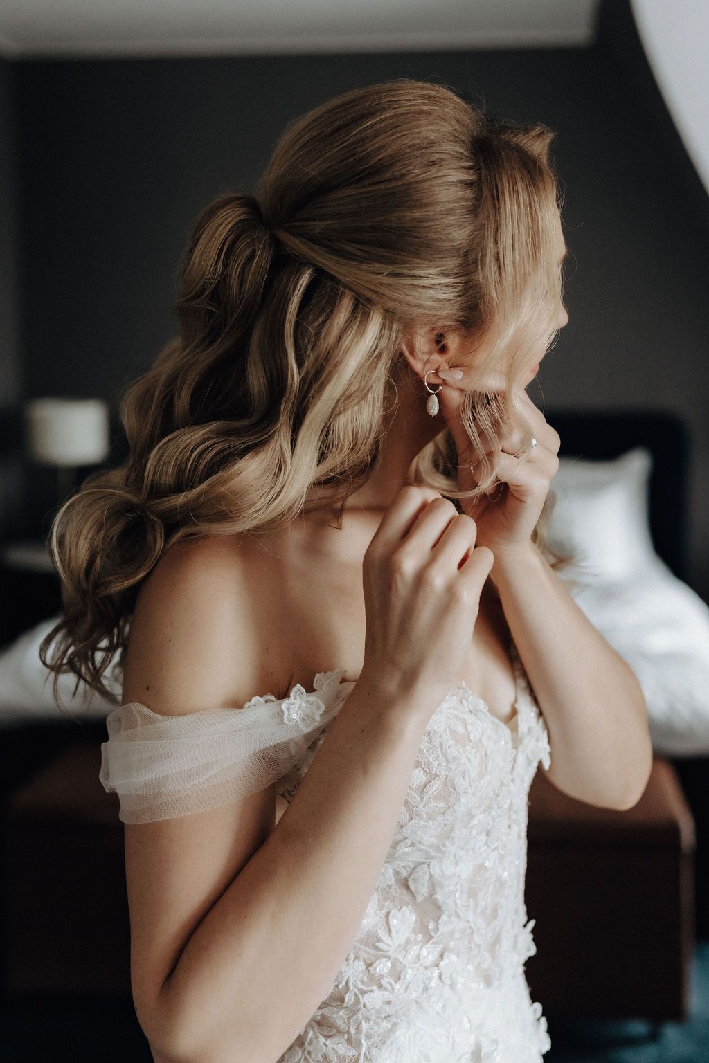Bride in floral lace wedding gown fastening drop pearl earrings before ceremony.