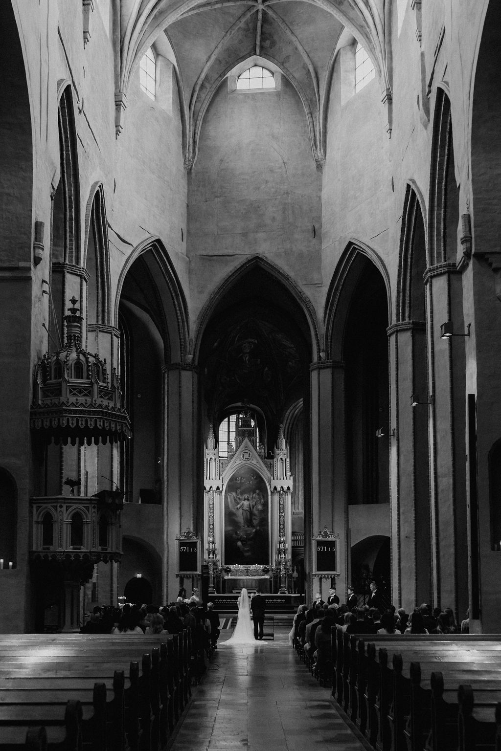 Black and white photo of bride walking down aisle of grand Gothic cathedral with vaulted ceilings.