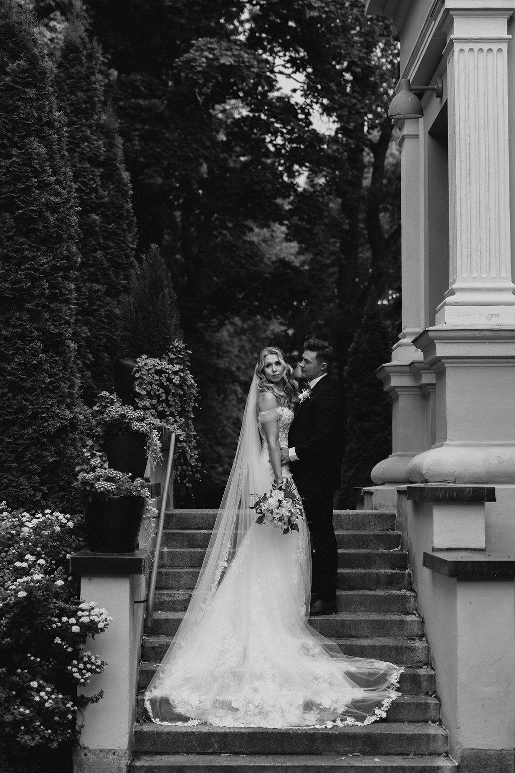 Black and white photo of bride and groom on ornate stone steps, bride in cathedral veil gown.