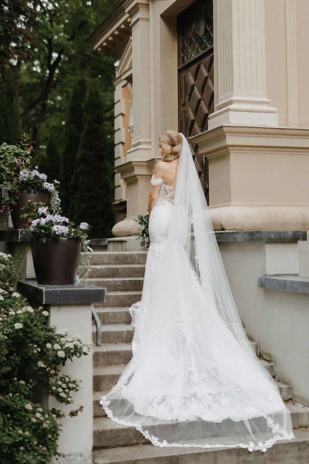 Bride in fitted lace wedding gown with long cathedral veil stands with back turned on ornate stone steps.