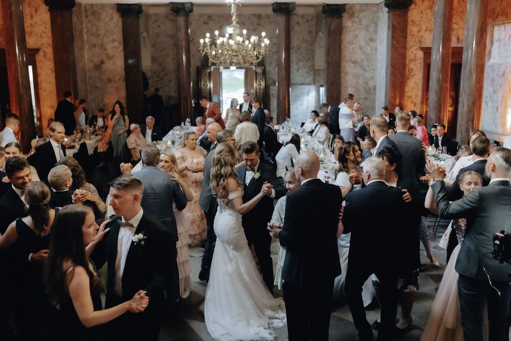 Bride and groom share first dance surrounded by guests in stunning chandelier-lit wedding venue.