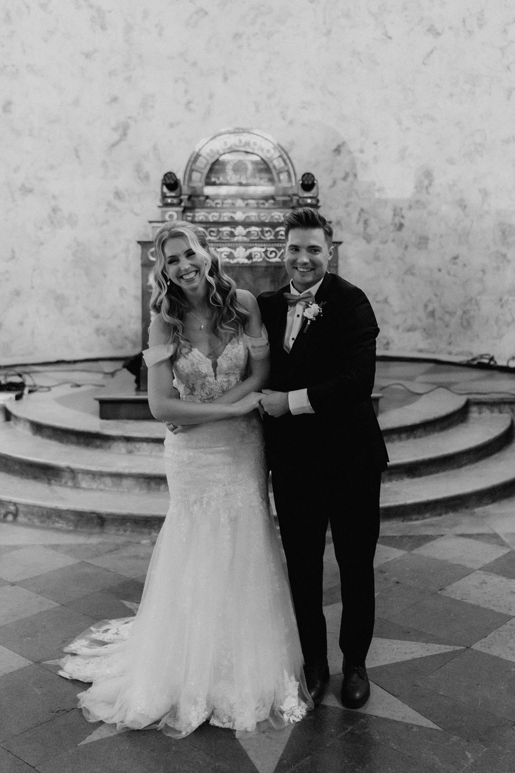 Smiling bride and groom pose together in black and white wedding portrait before ornate stone fountain.