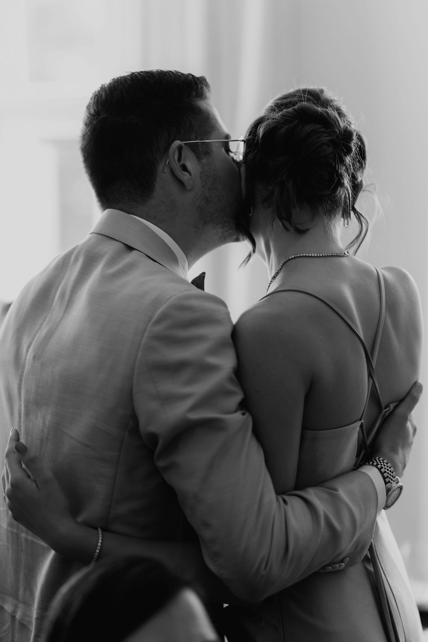 Black and white photo of man in suit kissing woman on head in tender wedding moment.