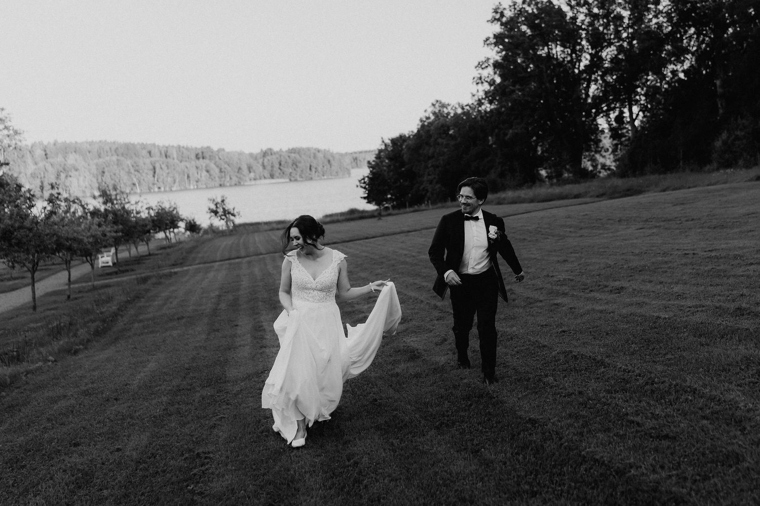 Black and white photo of bride and groom walking joyfully across a grassy lawn with a lake backdrop.