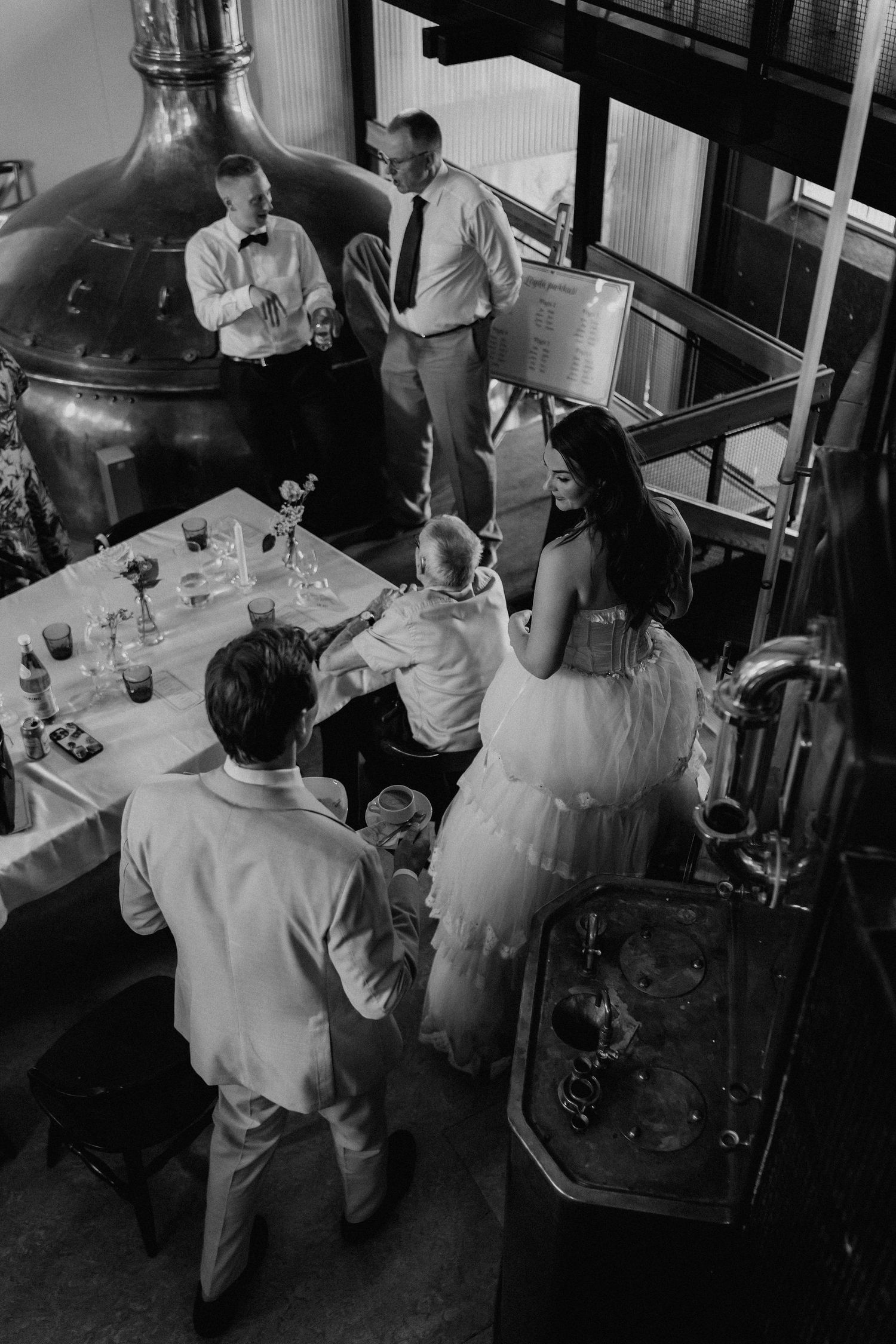 Black and white aerial view of bride and groom mingling with guests near copper brewery tanks.