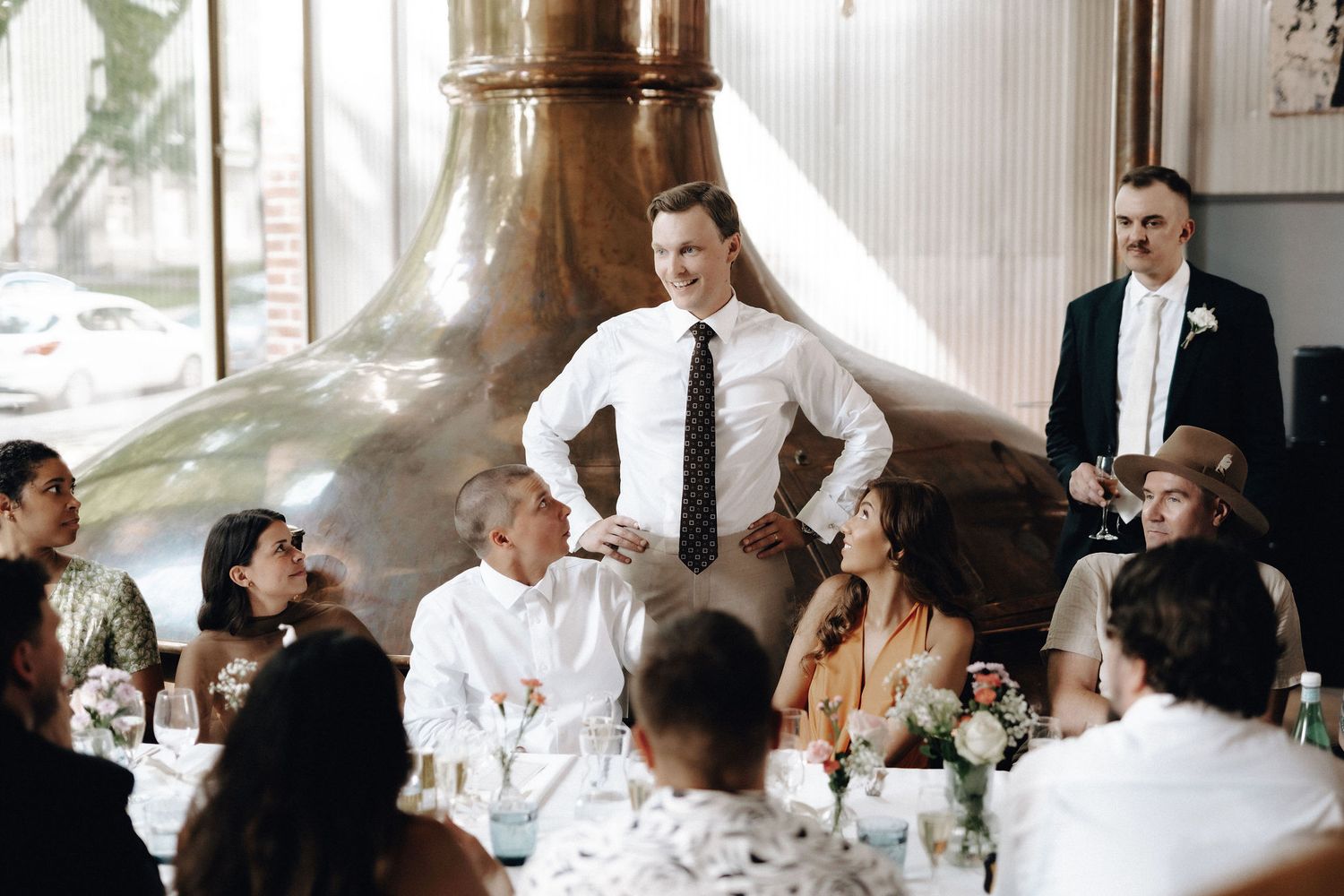 Groom in white shirt and tie laughing with wedding guests seated at decorated tables near a copper brewery tank.