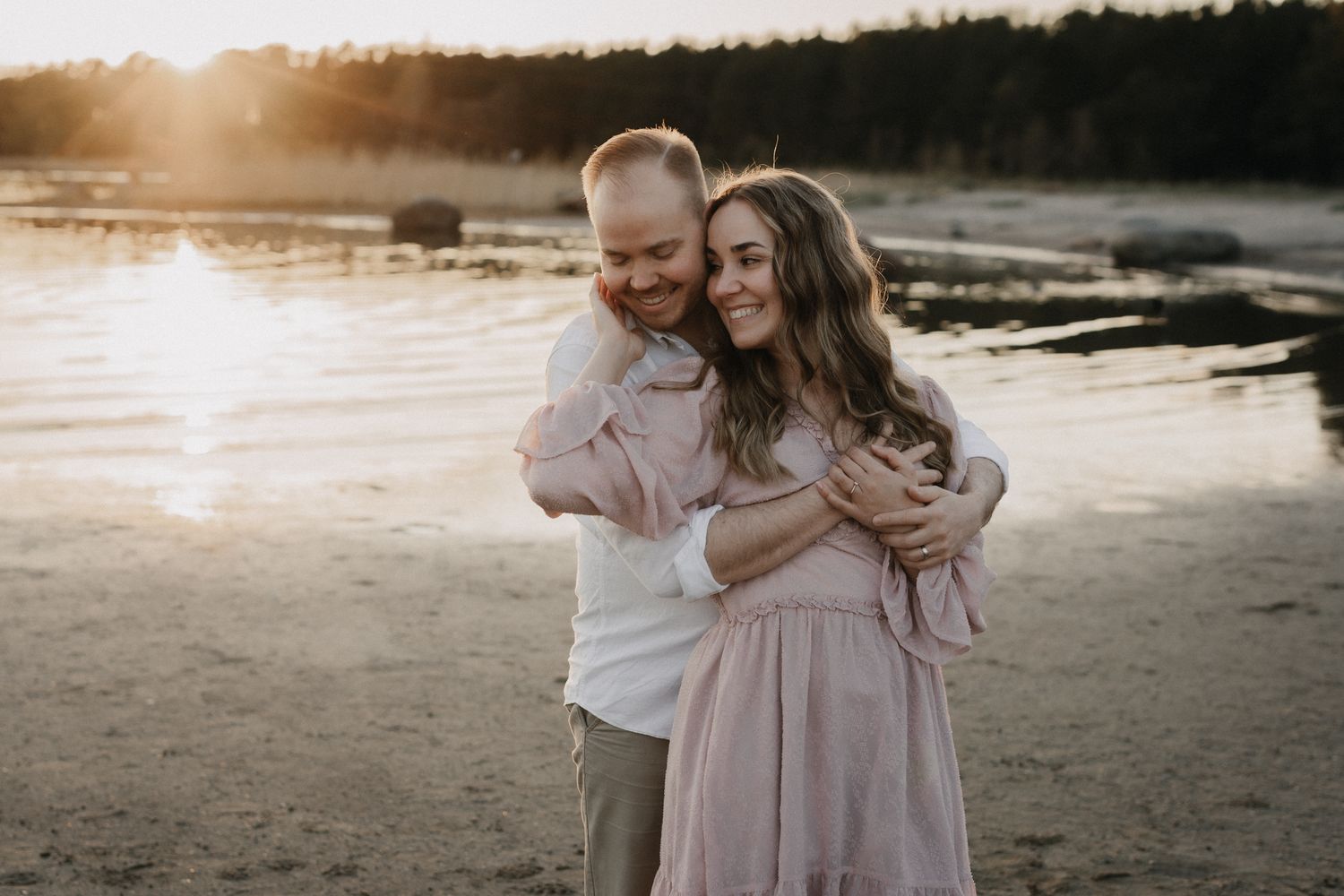 Couple shares a warm embrace on a sunlit beach, smiling together in soft golden hour light.