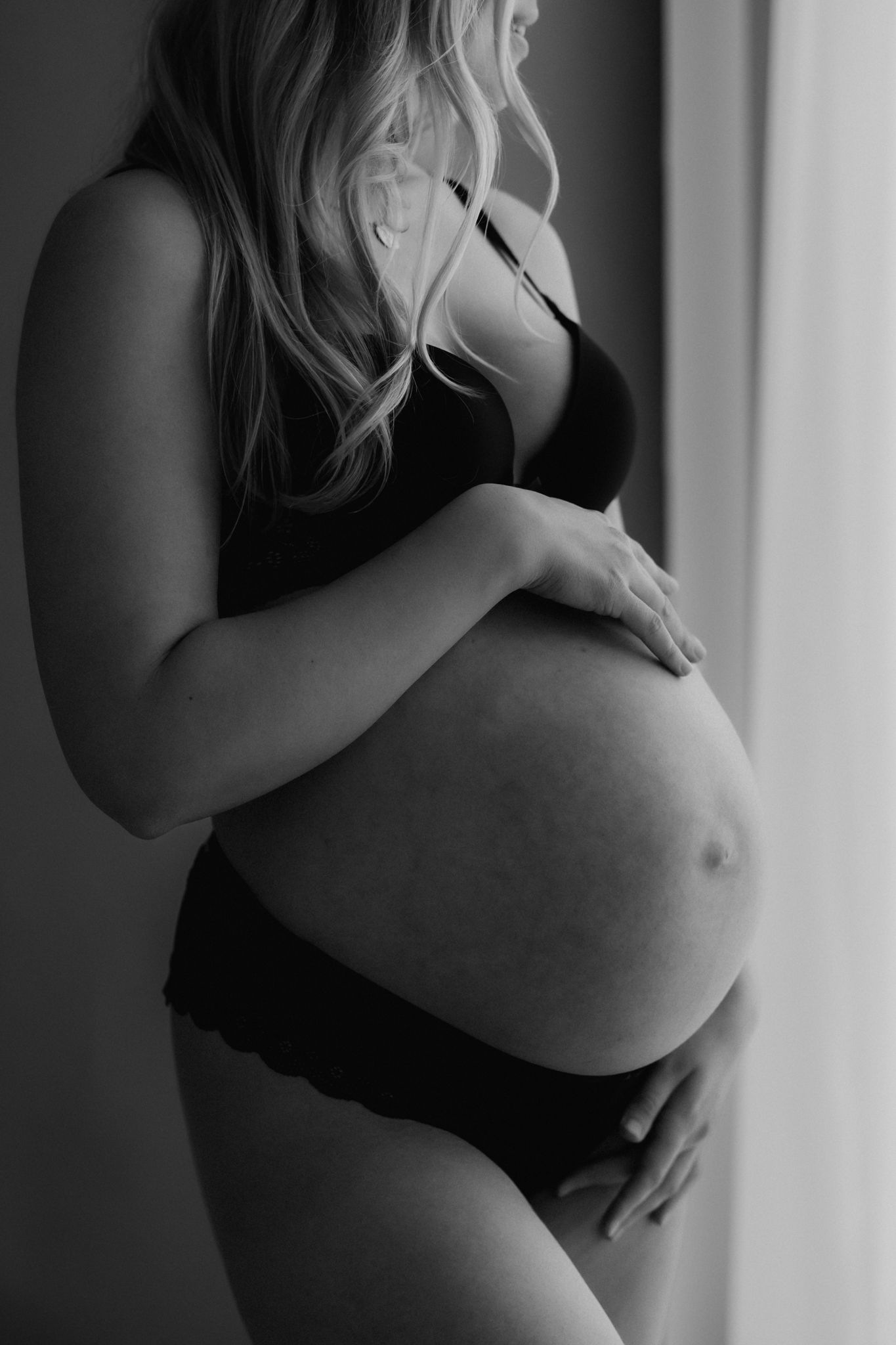 Pregnant woman in black lingerie holding her bare baby bump near window, black and white maternity photography.