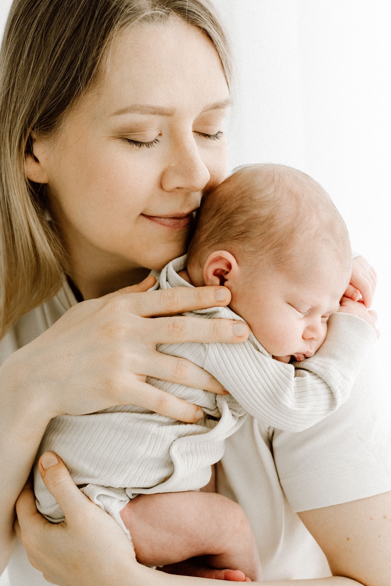 Mother with eyes closed tenderly holding sleeping newborn baby close to her cheek.