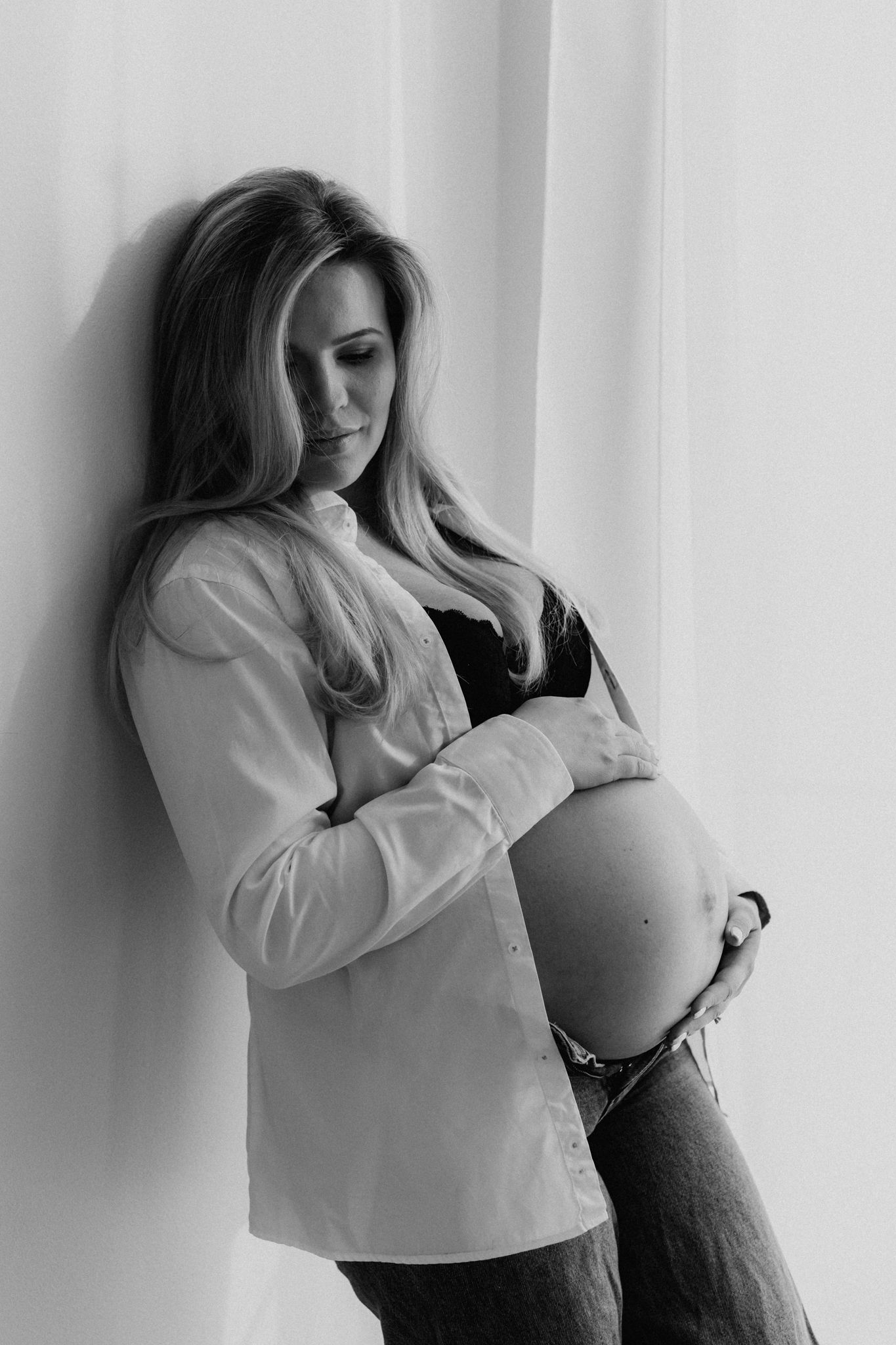 Black and white portrait of pregnant blonde woman in open white shirt holding bare bump.