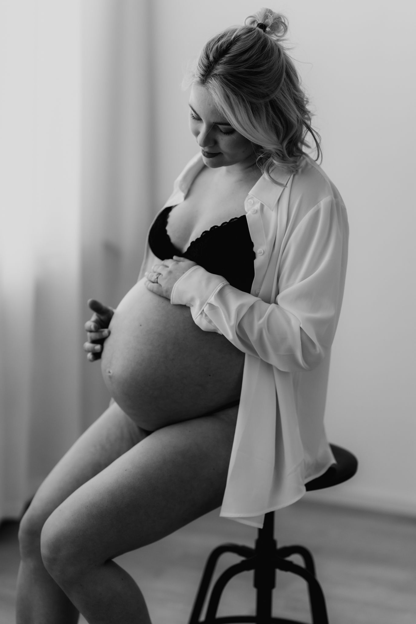 Black and white portrait of pregnant blonde woman in black bra and white shirt holding her belly.