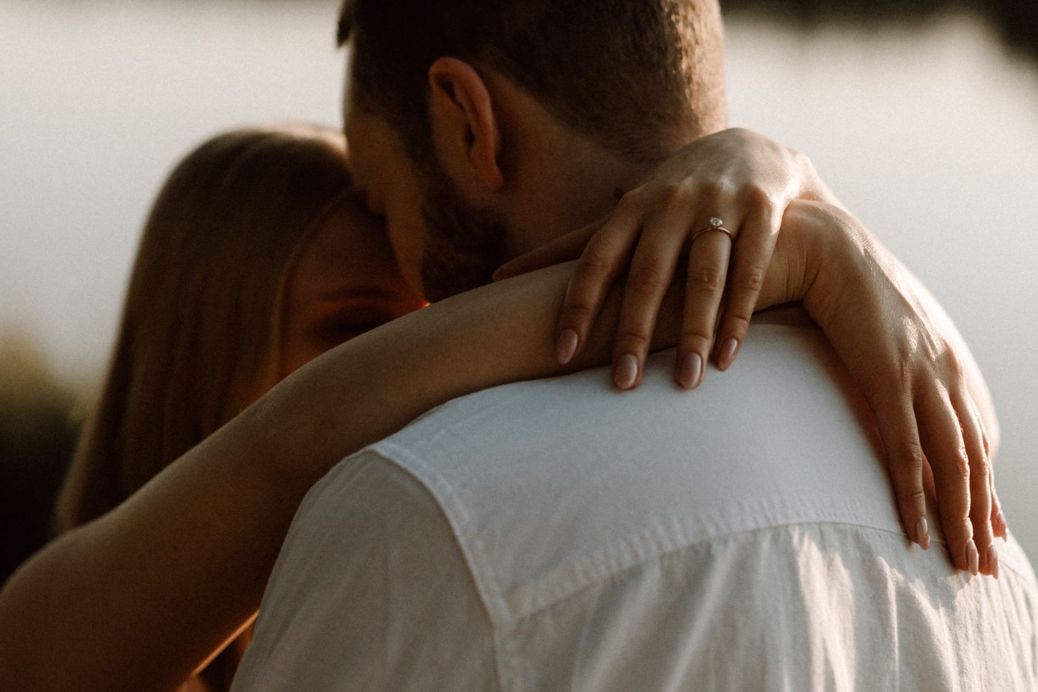 Close-up of a woman with an engagement ring embracing a man in a white shirt outdoors.