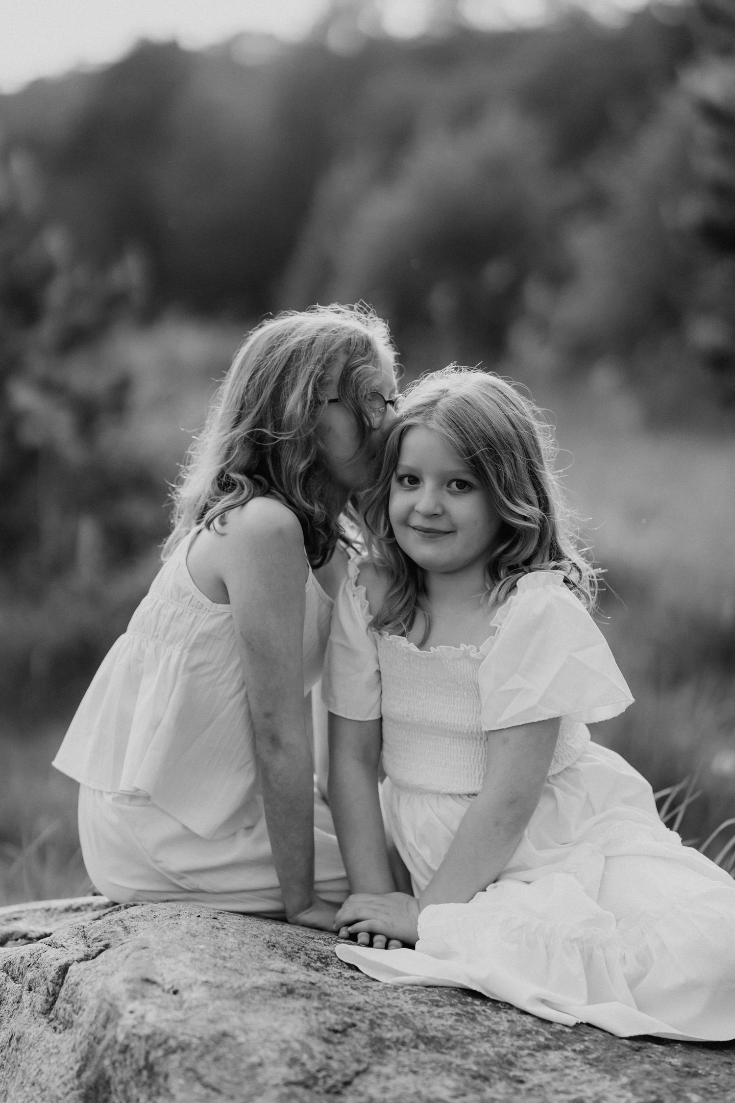 Two young sisters share a sweet moment on a rock outdoors in this black and white portrait photo.