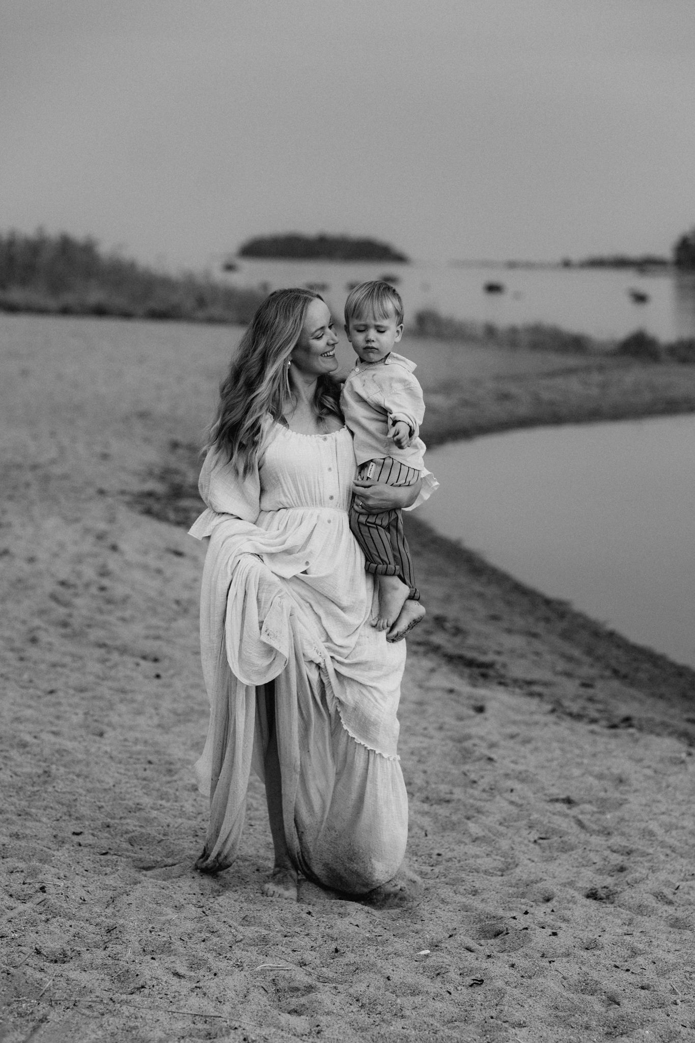 Black and white photo of a mother in a flowing white dress holding her toddler on a sandy beach.
