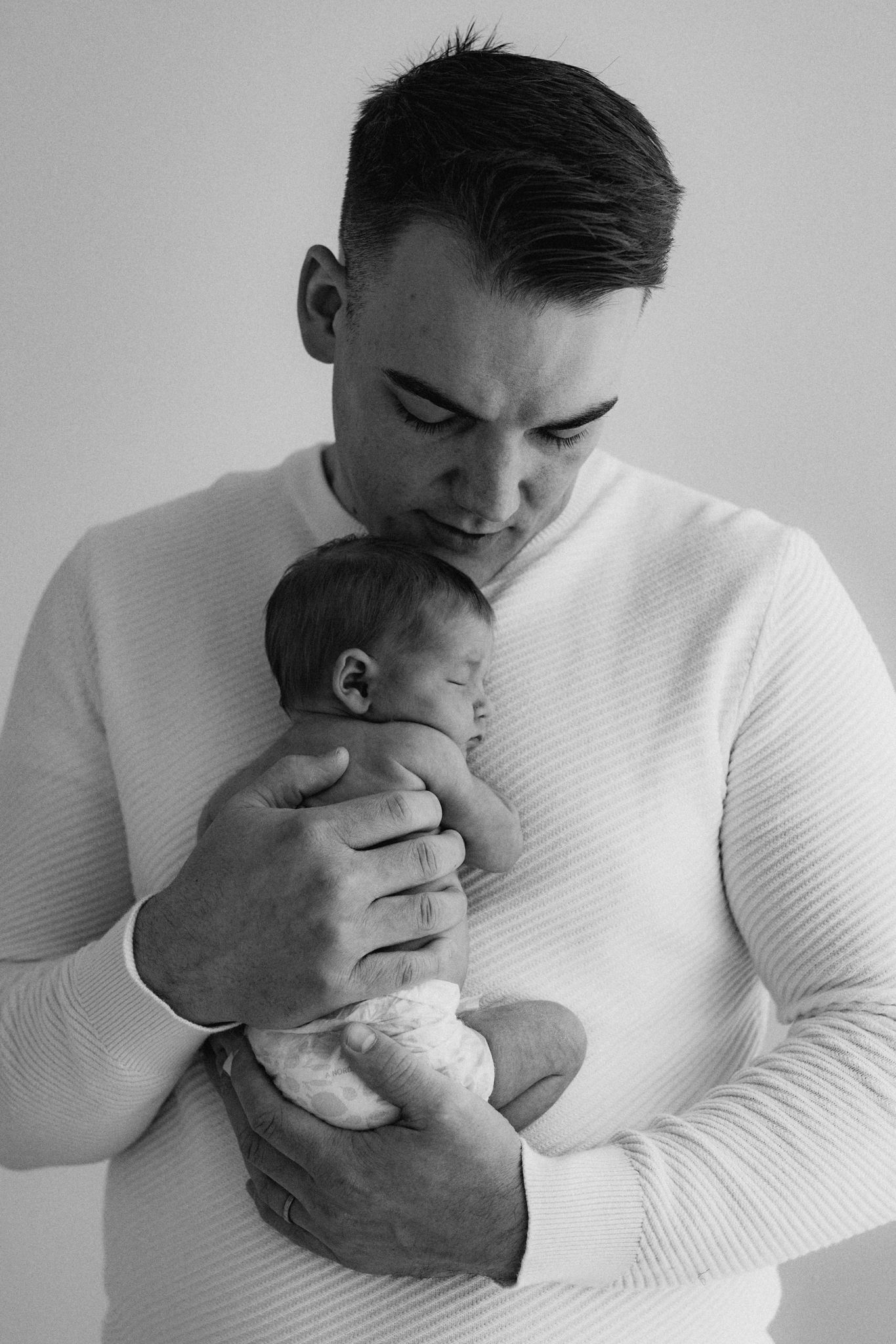 Black and white portrait of father in white sweater cradling sleeping newborn baby.