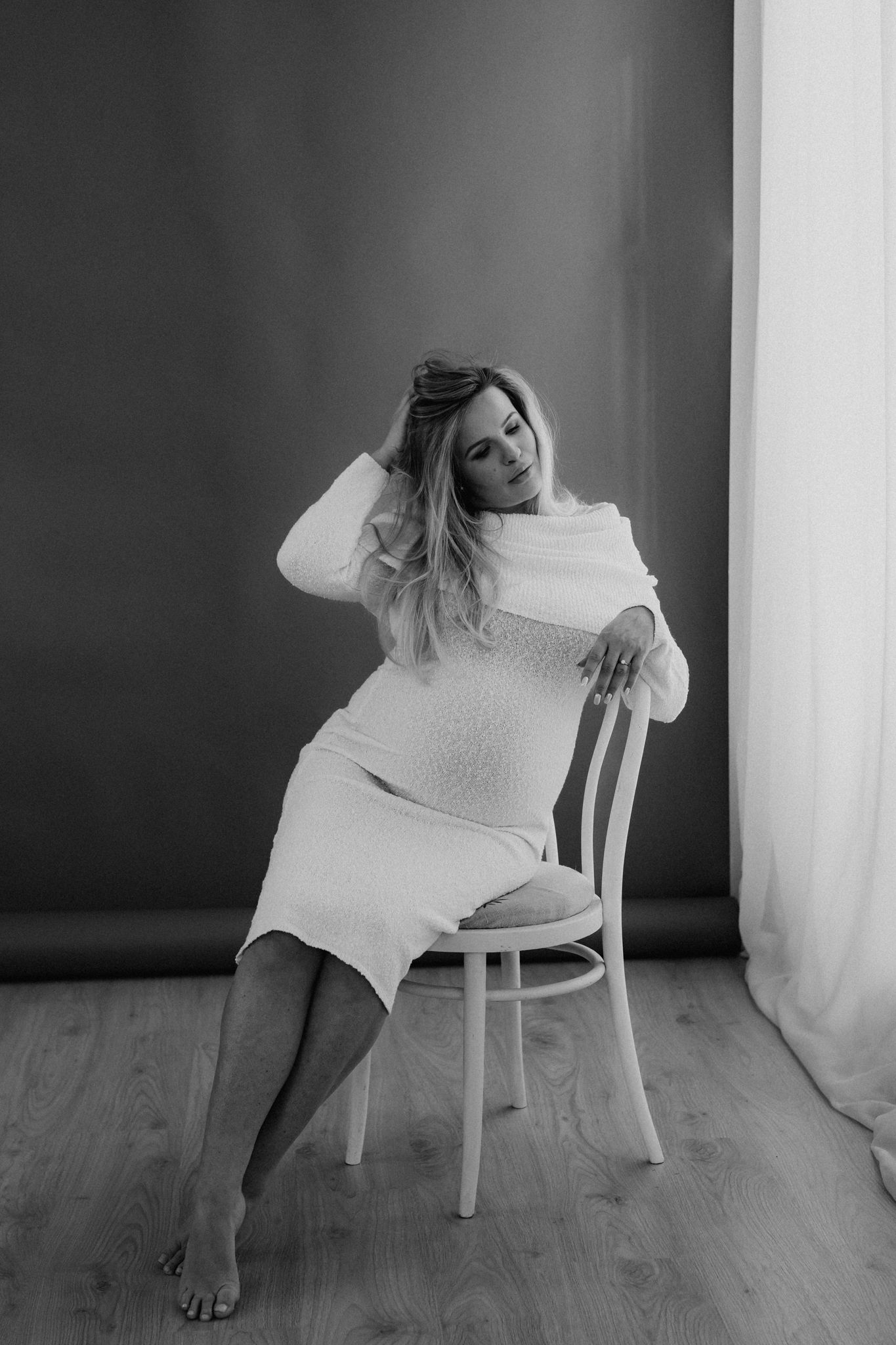 Black and white photo of pregnant woman in white dress seated on chair in studio setting.