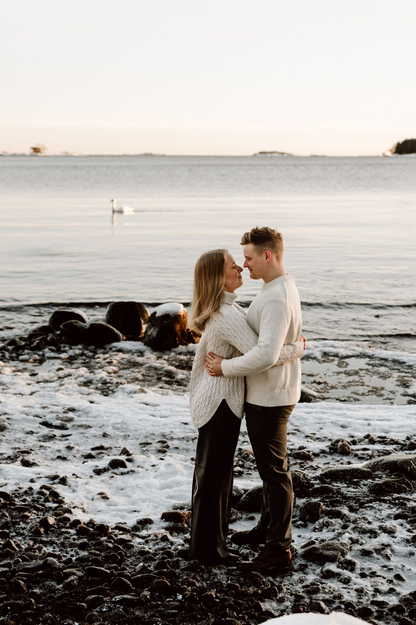 Couple embracing on a rocky snowy beach at sunset with a swan visible in the background.
