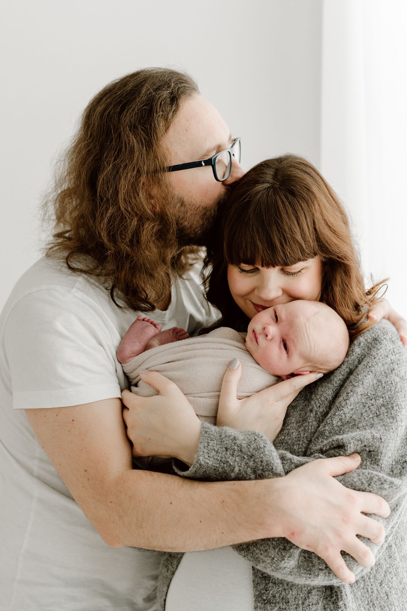 Loving parents with long hair kissing and cuddling their swaddled newborn baby together.