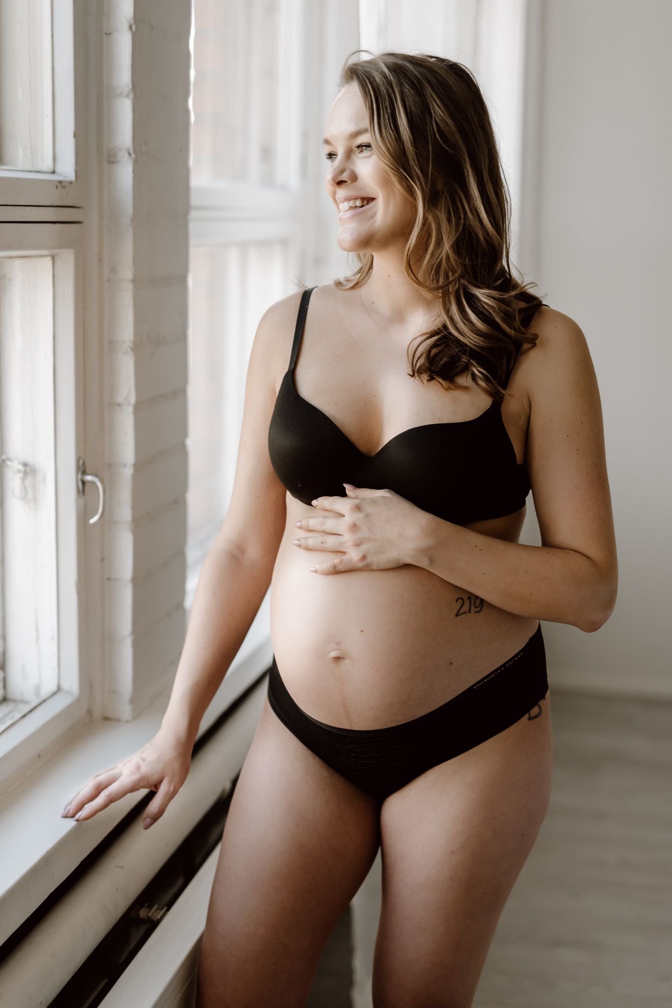 Smiling pregnant woman in black lingerie standing by window holding baby bump with tattoo.