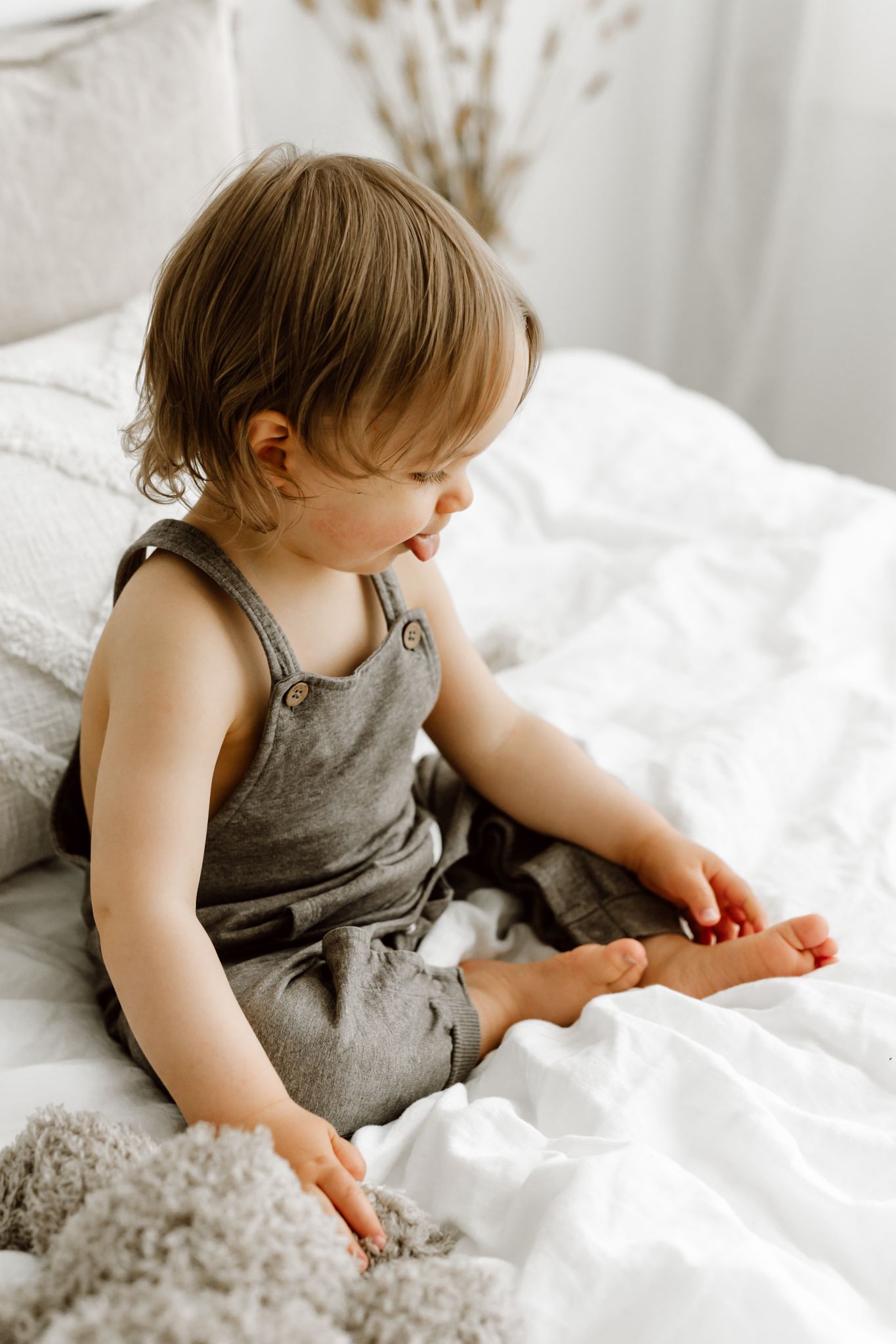Cute toddler in gray overalls sitting on white bed examining feet with stuffed animal nearby.
