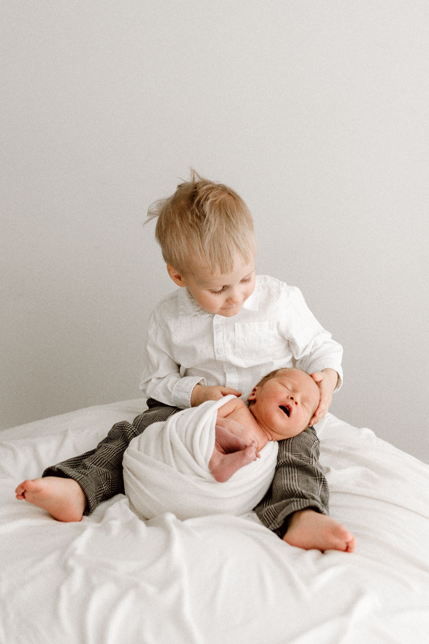 A young blonde toddler boy in a white shirt sits on a bed holding a yawning swaddled newborn.