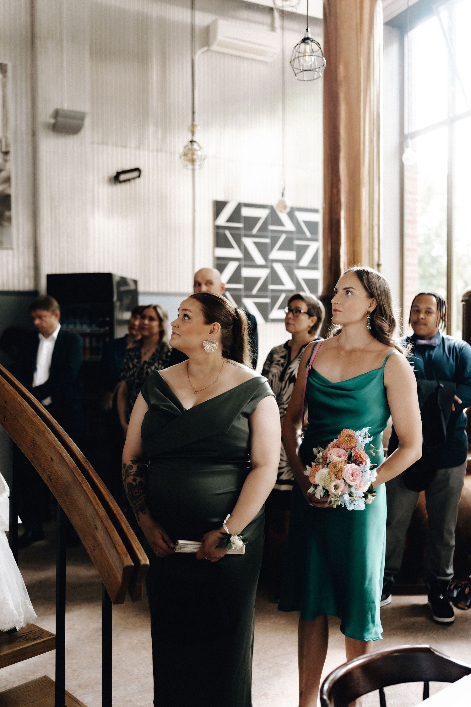 Two bridesmaids in emerald green satin dresses watch the wedding ceremony holding a floral bouquet.