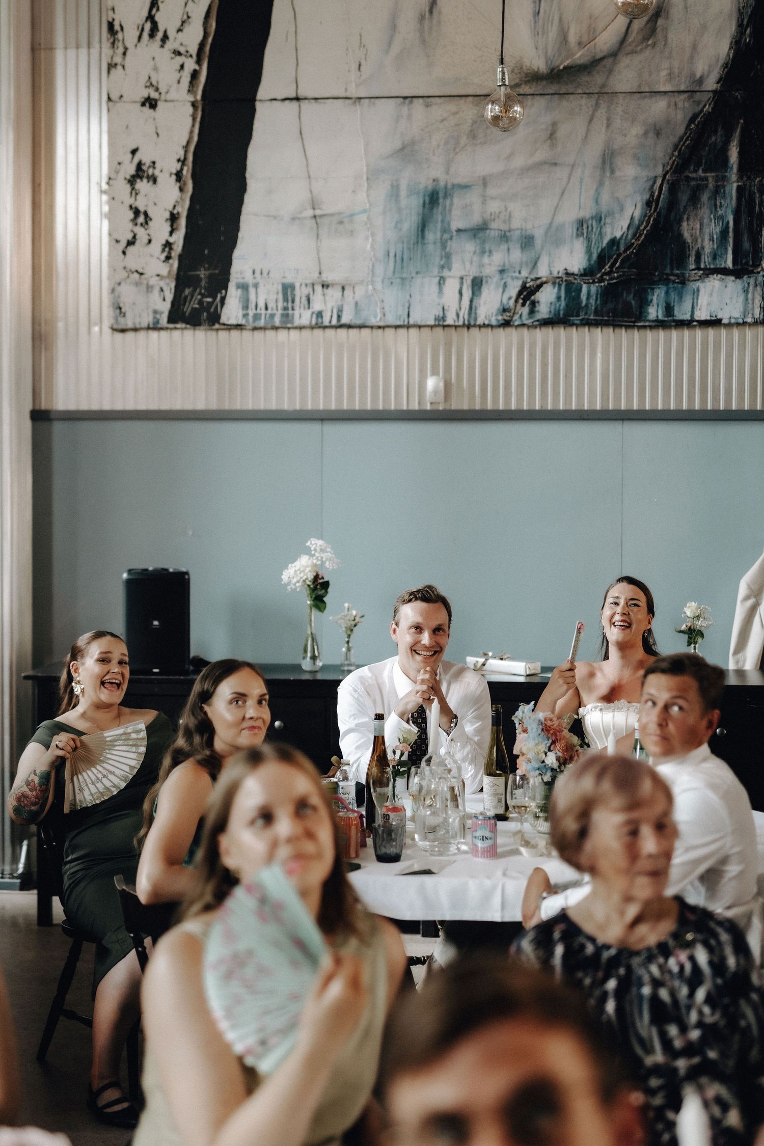 Wedding guests laughing and celebrating at reception tables beneath large abstract wall art.