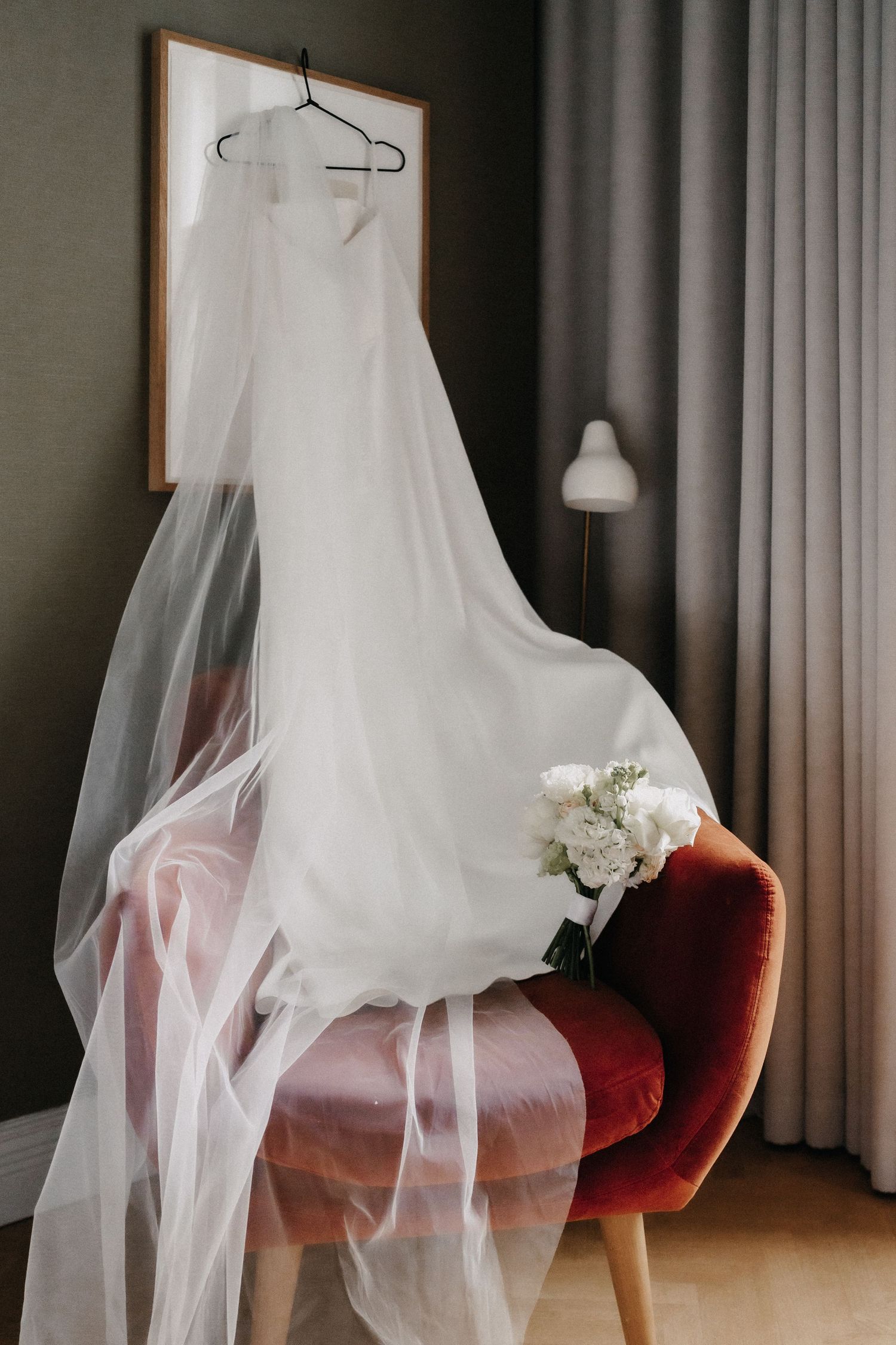 White wedding dress with flowing veil hanging over a red velvet chair beside a bridal bouquet.