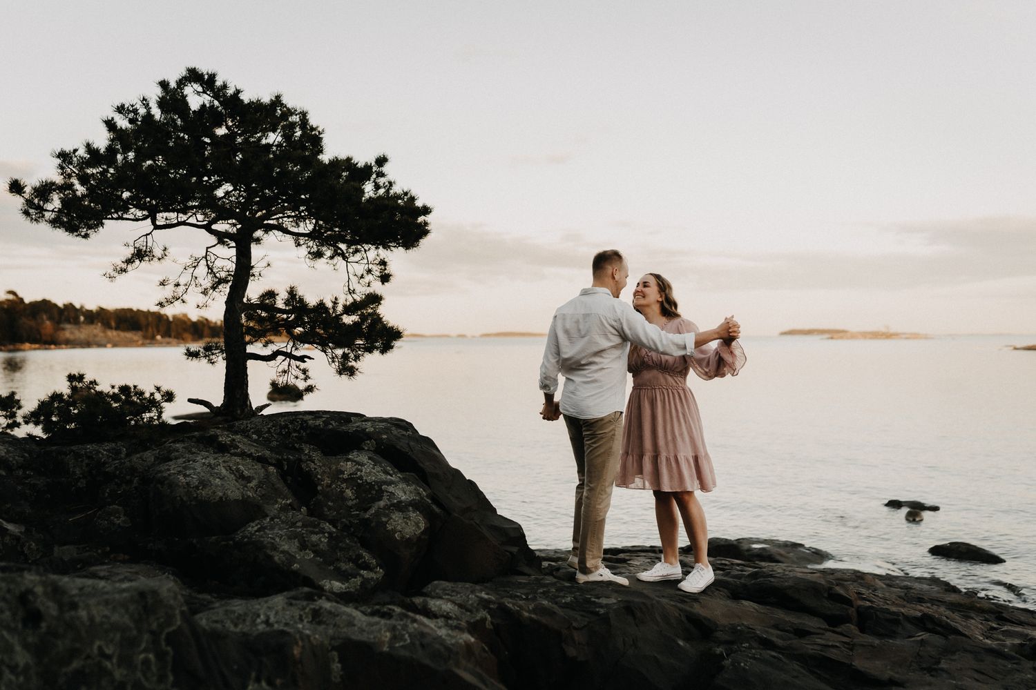 Pariskunta halailee kalliojyrkänteellä rauhallisen veden yllä auringonlaskussa yksinäisen männyn luona.