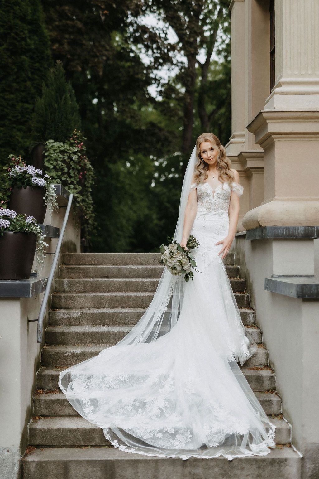 Bride in fitted lace wedding gown with flowing veil stands on stone staircase beside building.