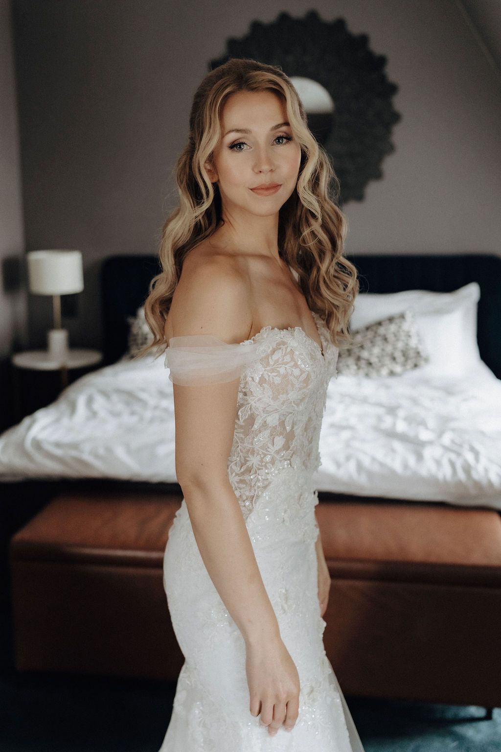 Beautiful bride in off-shoulder lace mermaid wedding dress posing in hotel room.